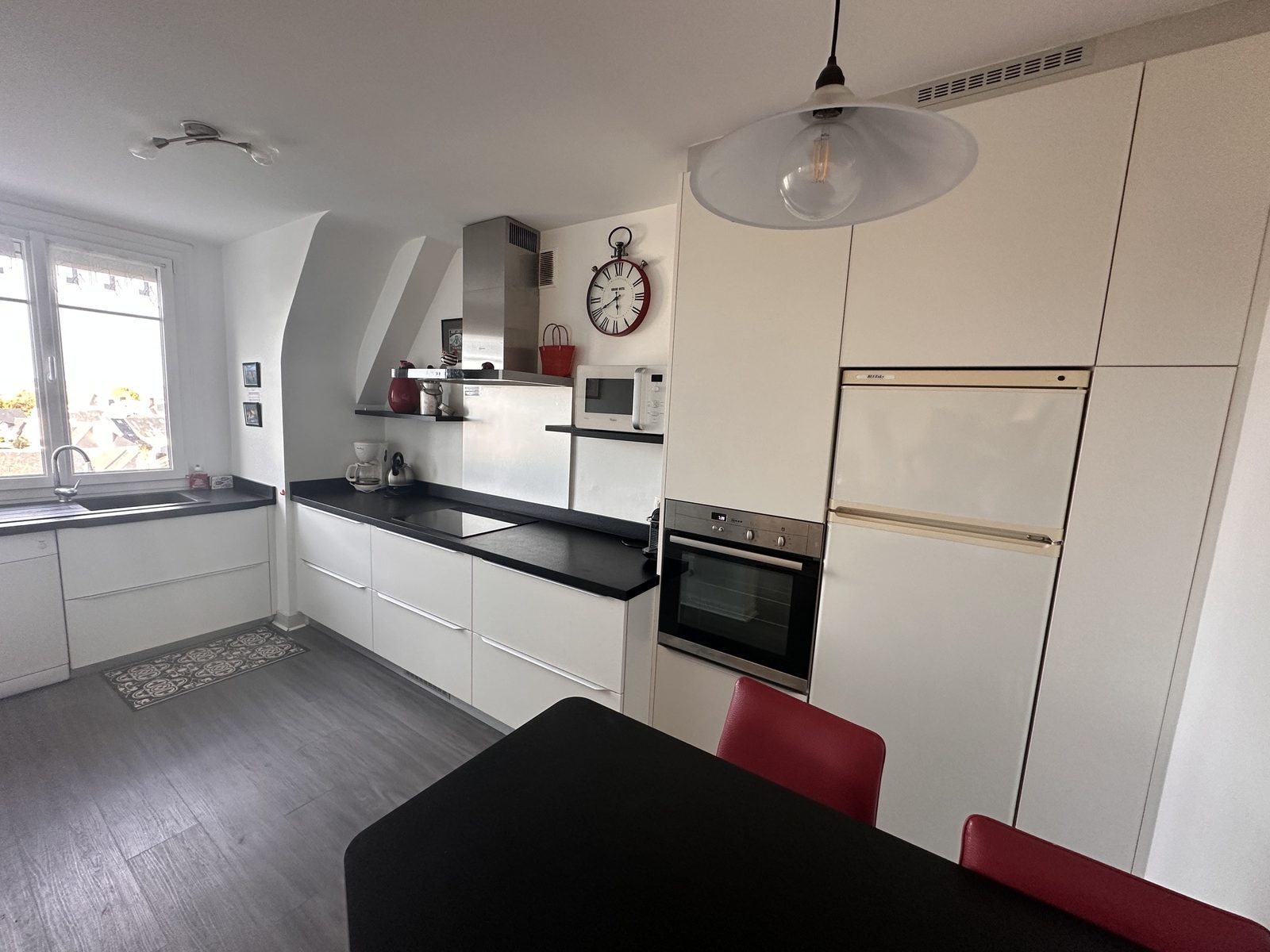 Cuisine en longueur, façades blanches et plan de travail noir : plaques à induction, four, micro-ondes, lave-vaisselle, grand réfrigérateur. Une fenêtre au-dessus de l'évier.