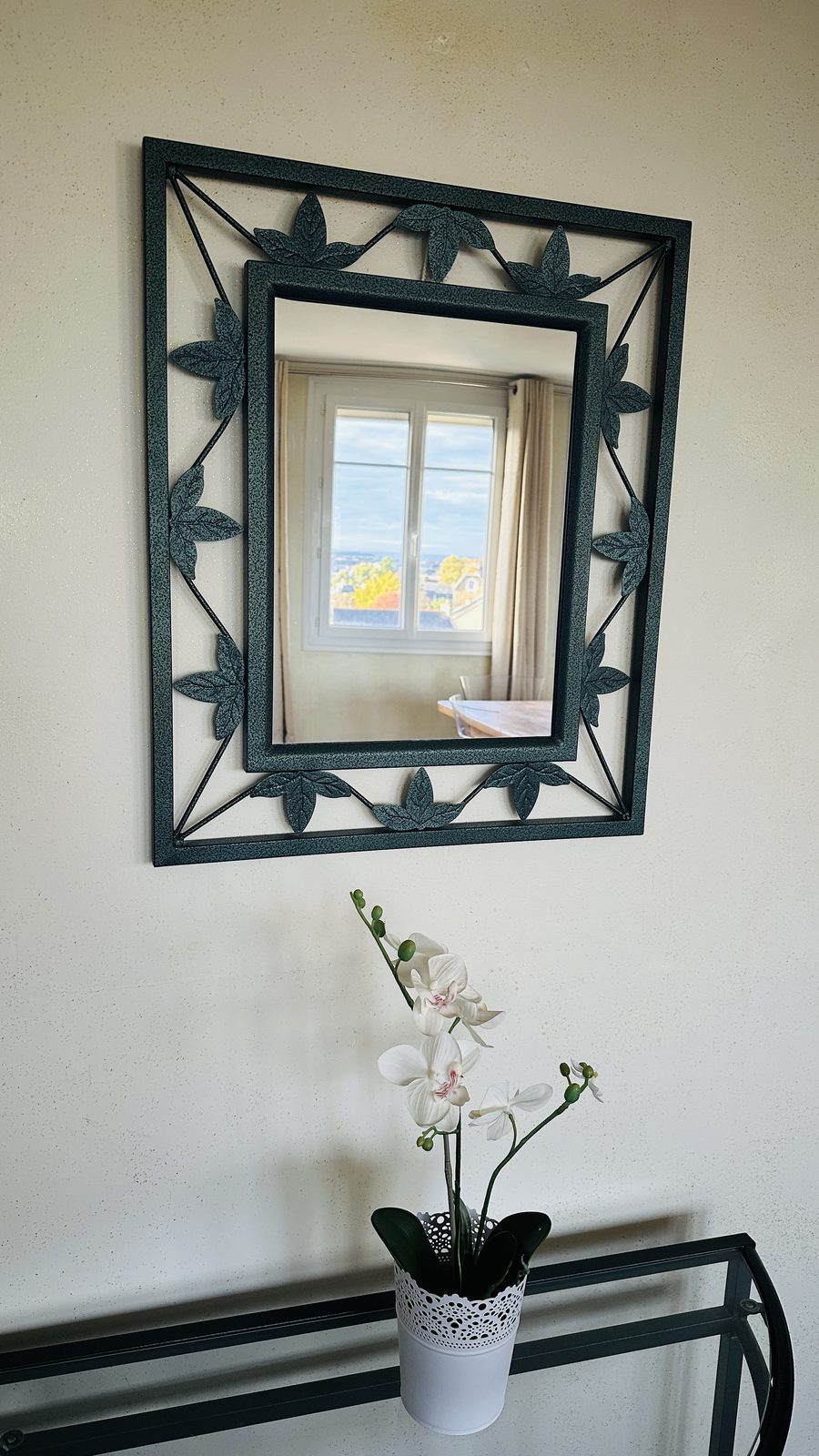 Dans l'entrée, un miroir au cadre de fer forgé au-dessus d'un guéridon, avec une orchidée blanche en pot.