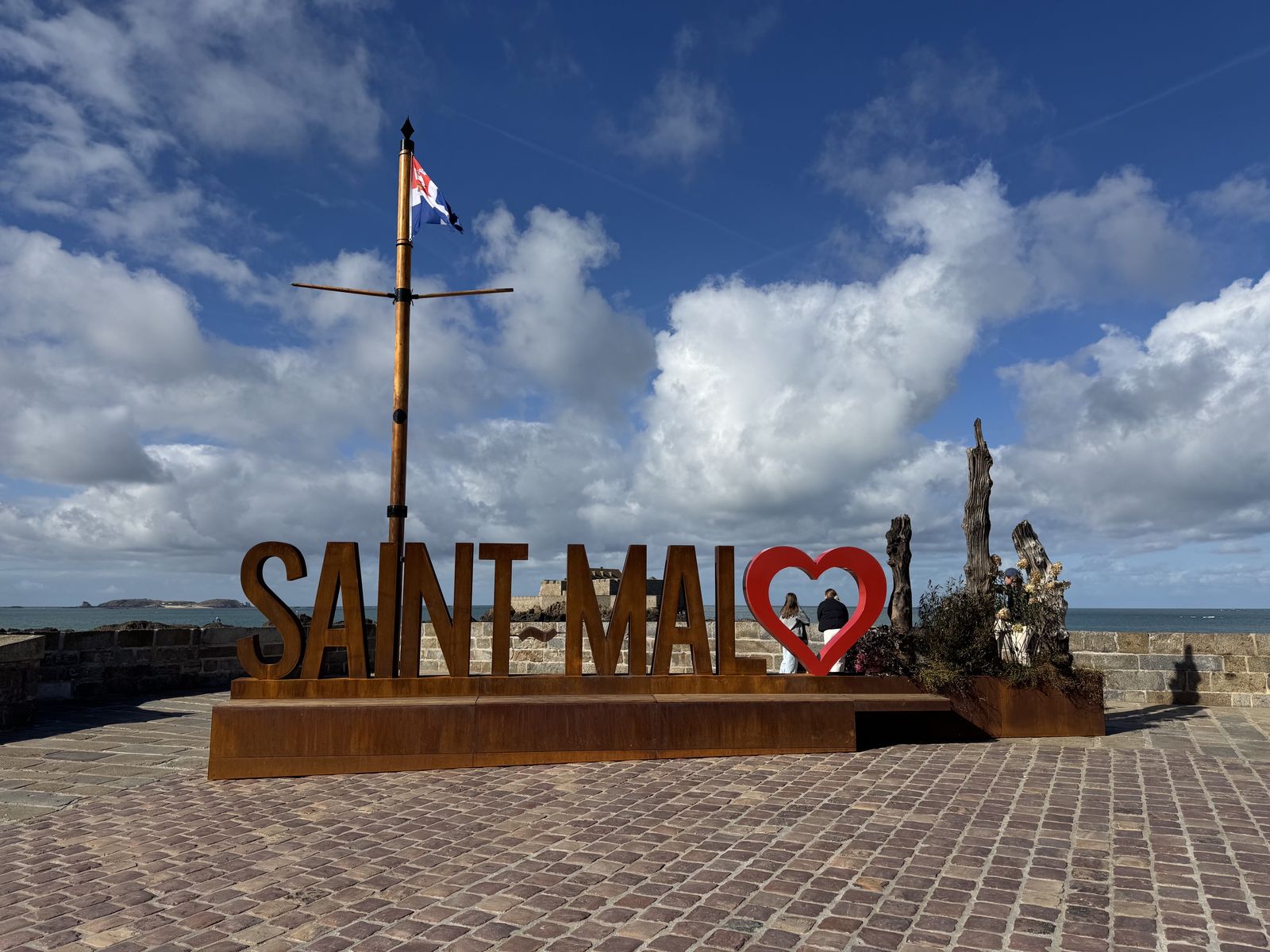 Sur le môle, les grandes lettres « Saint-Malo » en acier, le « o » en forme de cœur, devant la mer et un mât au drapeau malouin, sous un ciel de nuages.