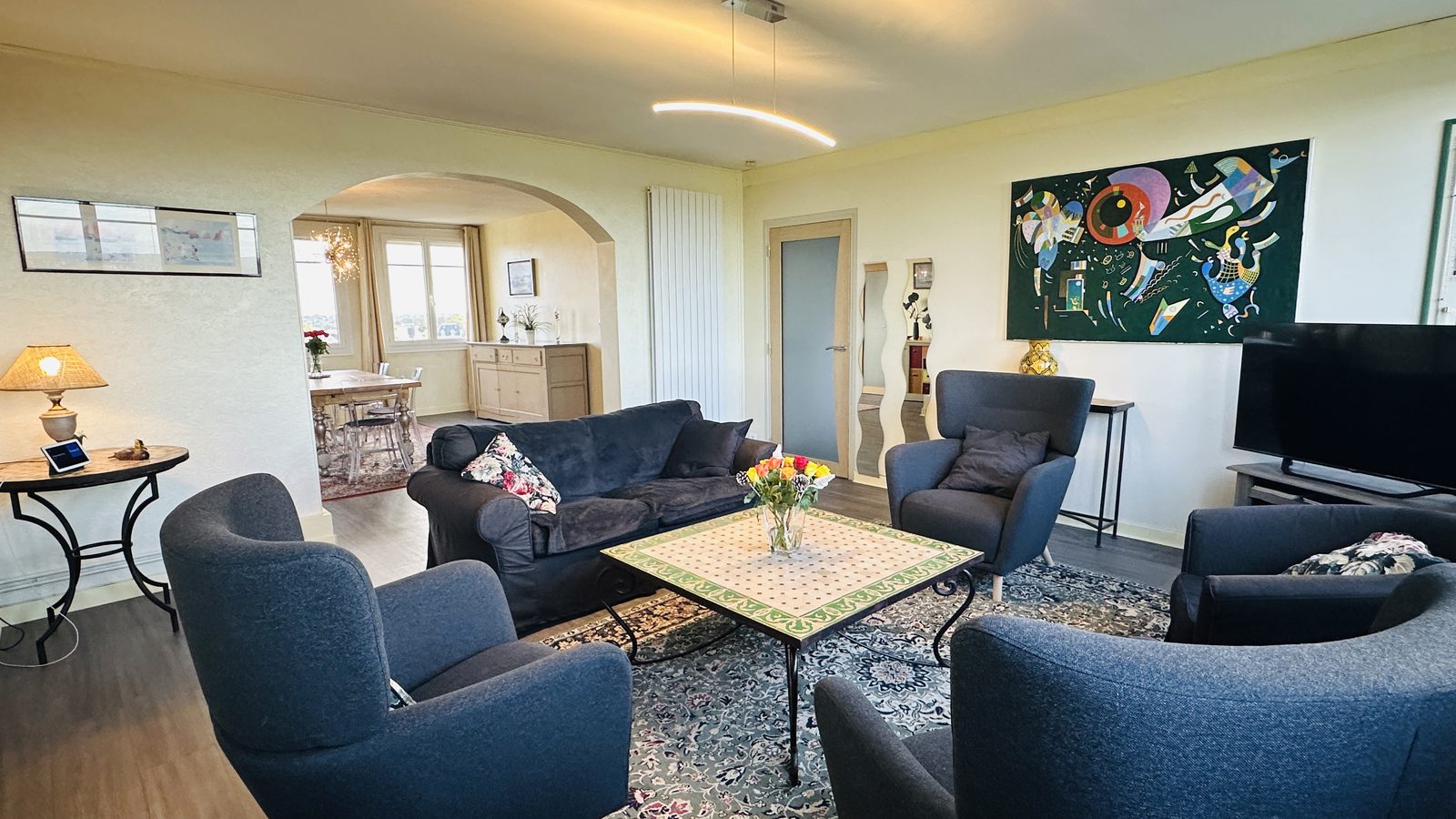 Le salon vu depuis les fenêtres : les fauteuils gris et le canapé autour de la table basse ; au fond, une arche ouvre sur le coin repas et sa table.