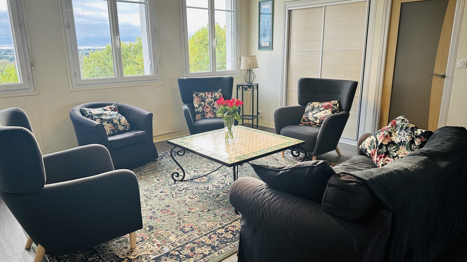 Salon lumineux : quatre fauteuils gris aux coussins fleuris et un canapé autour d'une table basse à plateau de faïence, sur un grand tapis à motifs ; trois fenêtres donnent sur la verdure et, au loin, la mer.