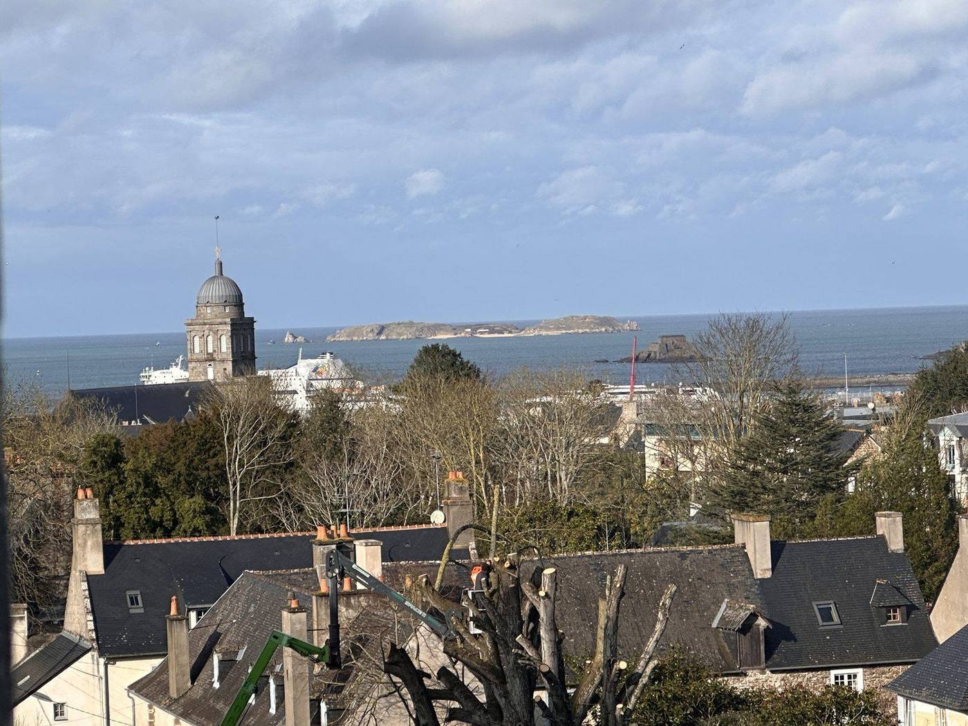 Par la fenêtre : au premier plan les toits d'ardoise et leurs cheminées, puis le clocher à dôme gris, un ferry blanc à quai, la mer, et à l'horizon des îlots rocheux et un fort.